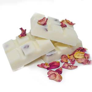 Three white chocolate bars with pink-purple inclusions, placed on a white background, are stacked slightly askew. Dried rose petals are scattered around them, adding a decorative touch.
