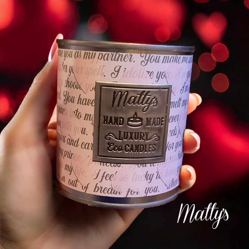 A person holds a candle labeled "Matty's Handmade Luxury Eco Candles" against a background filled with red glowing heart shapes. The candle has script text wrapping around it.