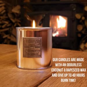 A lit metallic candle sits on a wooden table. A fireplace with stacked logs is blurred in the background. Text reads: "Our candles are made with an odourless coconut & rapeseed wax and give up to 40 hours burn time!.