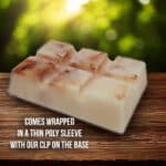 A handcrafted soap block with a marbled texture rests on a wooden surface. The background is blurred greenery. Text on the image reads: "Comes wrapped in a thin poly sleeve with our CLP on the base.