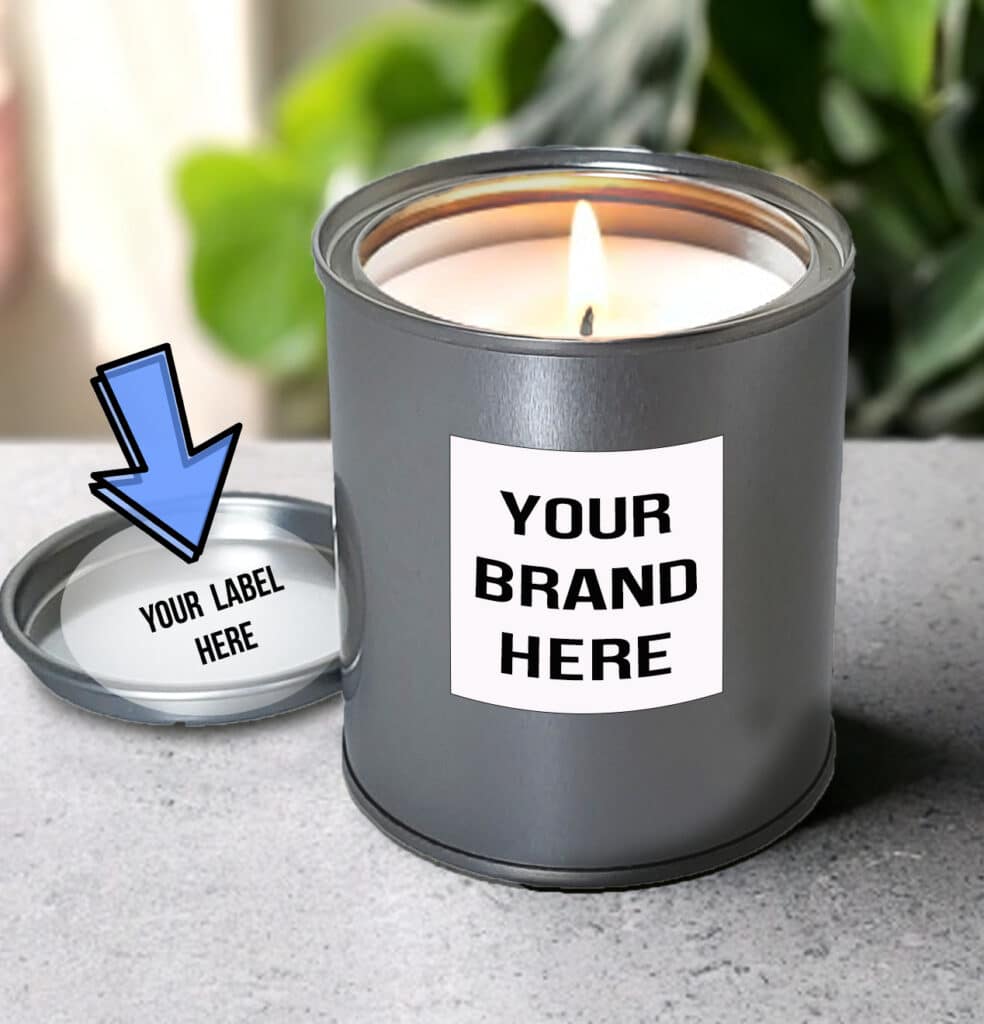 White Label Candle in a silver tin with "YOUR BRAND HERE" on the label showcases our White Label Candles. The lid, placed beside it, bears "YOUR LABEL HERE" and an illustrated blue arrow pointing to it. Green leaves blur softly in the background, adding a touch of nature's elegance.