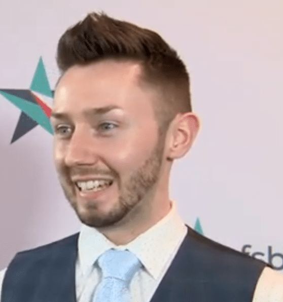 A man with short brown hair and a trimmed beard, wearing a white shirt, blue tie, and dark vest, smiling in front of a backdrop with a star graphic and partial text.