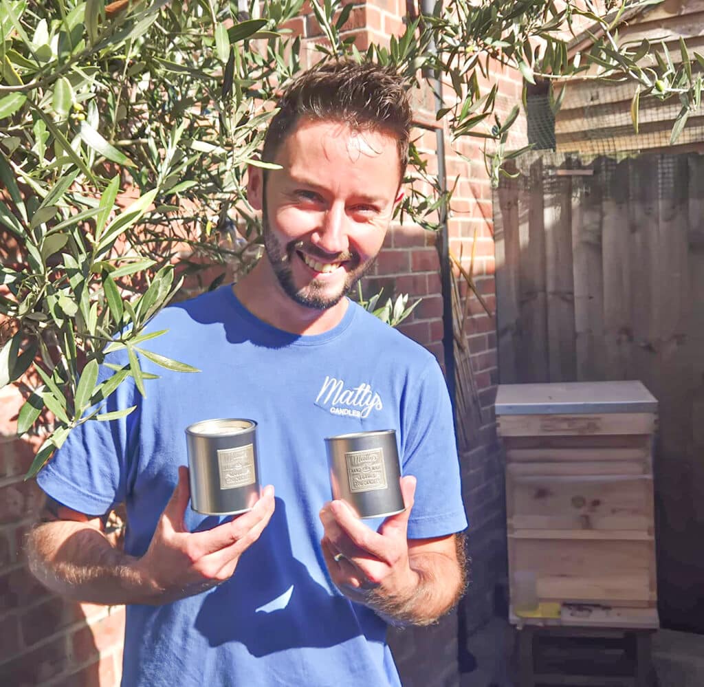 A smiling man in a blue "Matty’s" shirt stands outdoors near a beehive, holding two metal tins, surrounded by green foliage and a brick wall in bright sunlight.
