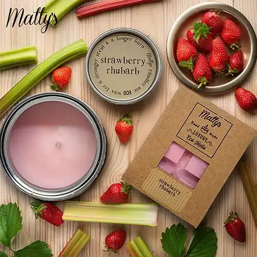 A pink candle and a box of candles and wax melts, labeled “Strawberry Rhubarb,” are arranged with fresh strawberries and rhubarb stalks on a wooden surface. The brand "Matty’s" is visible on the products and in the image.