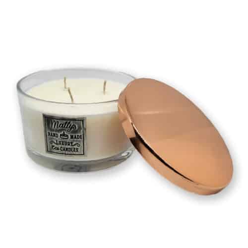 A white, three-wick candle in a clear glass container with a metallic label that reads "Matty's Handmade Luxury Eco Candles." The candle's round, shiny rose-gold lid is resting beside it.