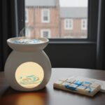 A lit wax melt burner emits steam on a wooden table near a window with raindrops. White wax melts with blue swirls and packaging sit beside it. Houses are visible outside the rainy window.